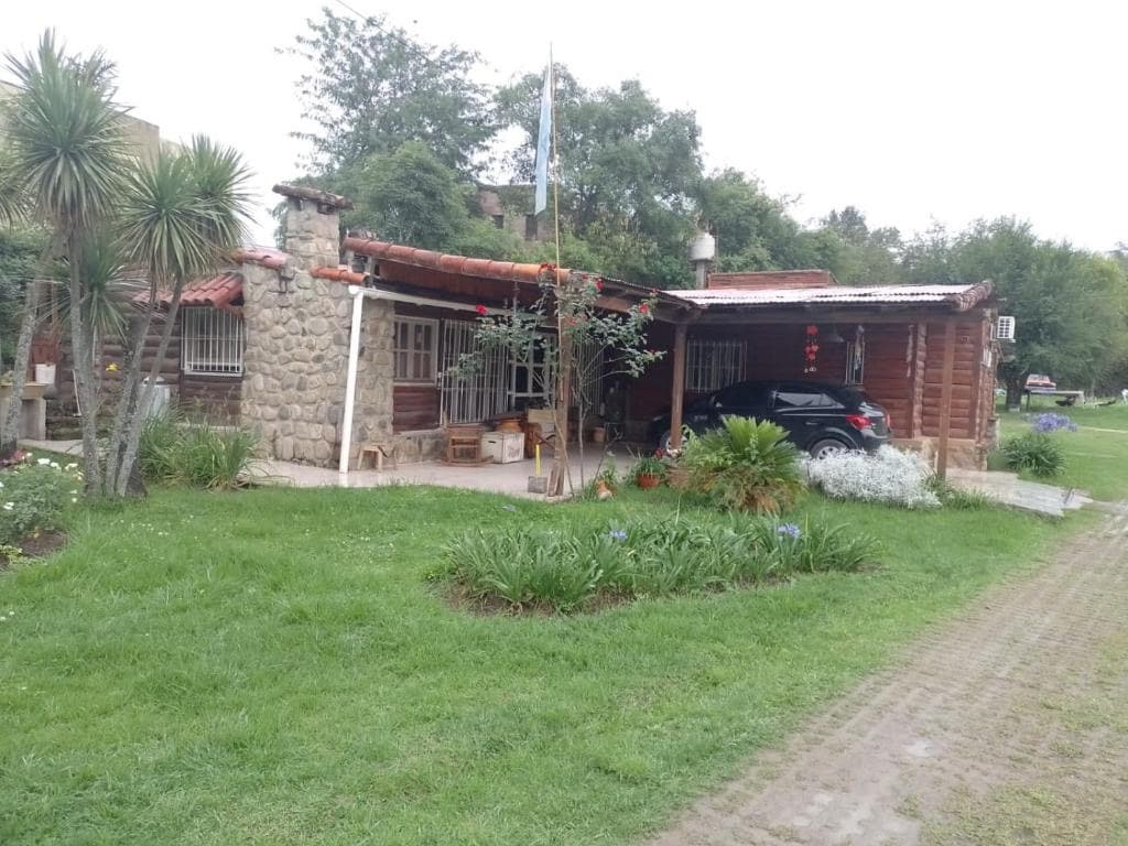 A charming rustic villa is surrounded by lush greenery and flowering plants. The structure features a combination of stone and wooden exterior. A car is parked nearby, and a dirt path leads to the entrance, emphasizing a tranquil connection with nature.