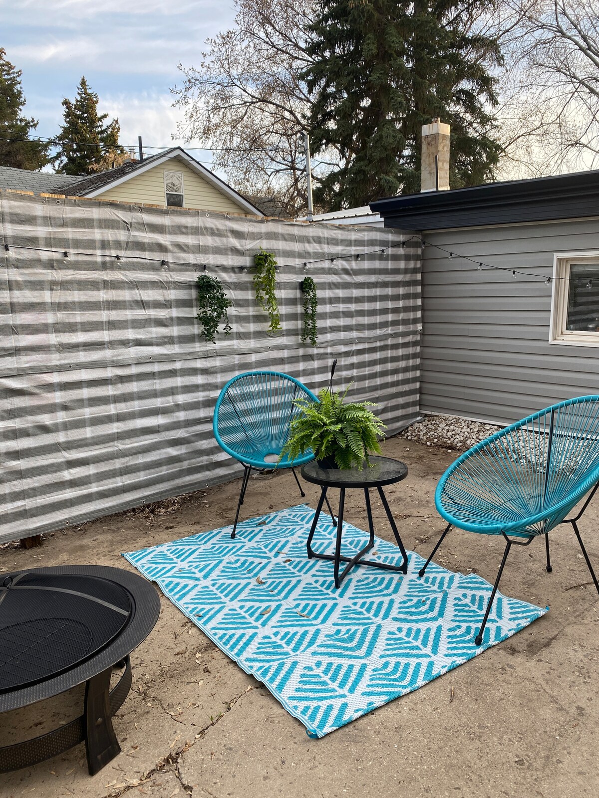 An outdoor seating area is presented with two teal acapulco chairs and a small black table, adorned with a lush green plant. A patterned blue and white rug covers the concrete floor, while a fire pit is placed nearby. Gray siding provides a neutral backdrop.