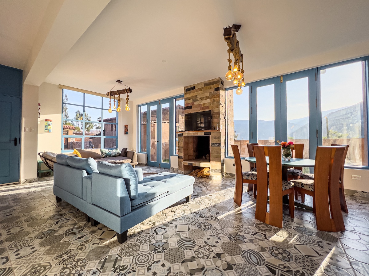 A spacious living area is highlighted by large glass windows, offering views of the surrounding landscape. A comfortable blue sofa faces a stone fireplace, while a wooden dining table accommodates several chairs. Unique patterned tiles adorn the floor, adding character to the space.