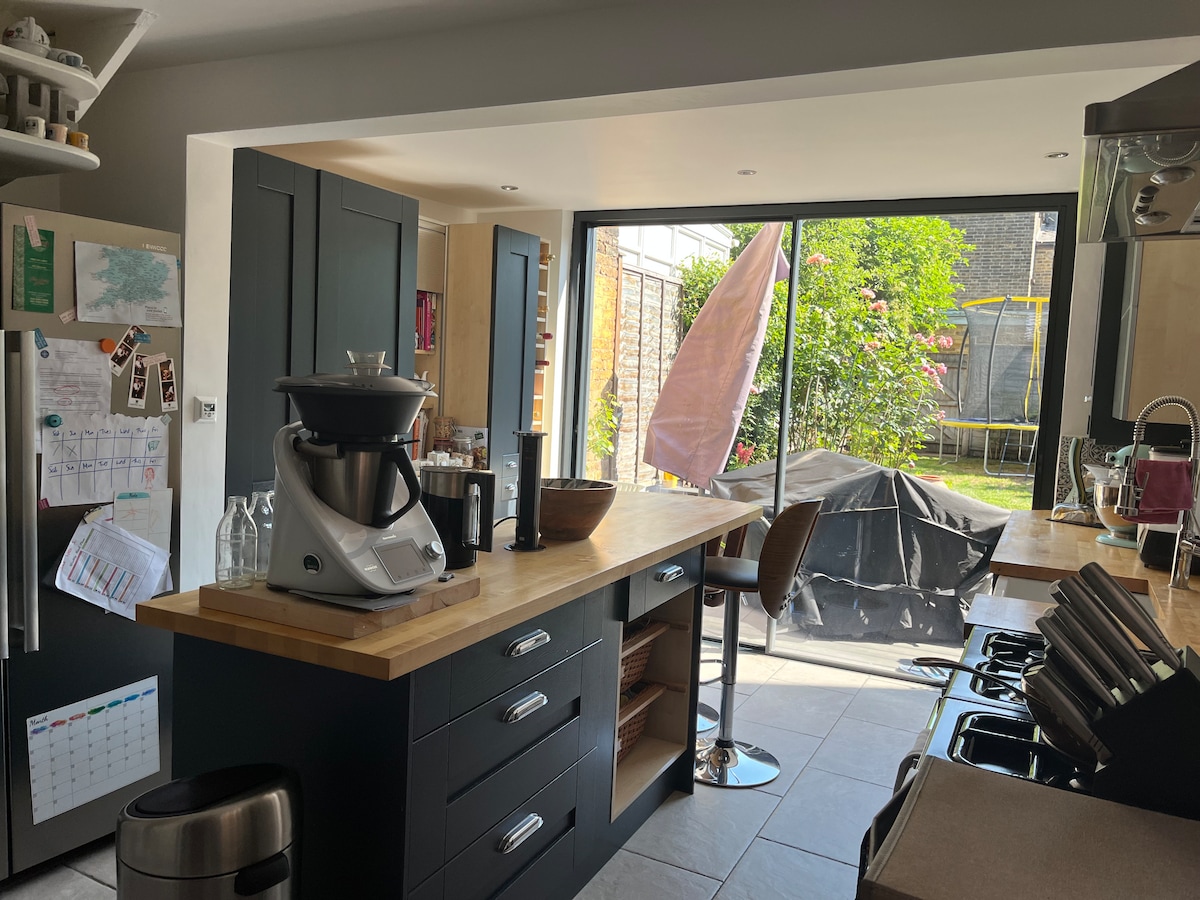 The kitchen features a modern design with dark cabinetry and a wooden countertop. A view into the garden is visible through large glass doors. A kitchen island with a bar stool is positioned near cooking appliances, while a blender and glassware adorn the countertop.