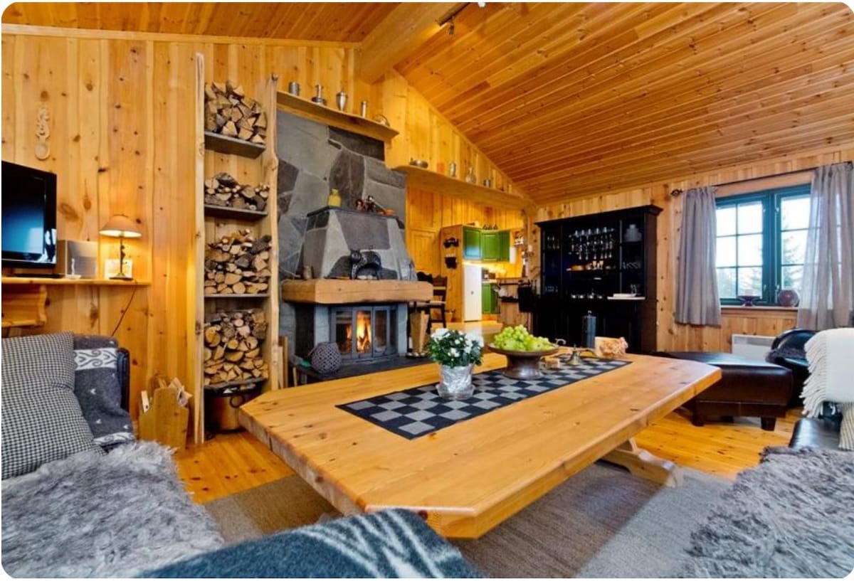 A spacious living area features warm wooden walls and a high ceiling. A large wooden table occupies the center, while a rustic stone fireplace is flanked by neatly stacked firewood. Light filters through large windows, complementing the cozy atmosphere created by the decor.
