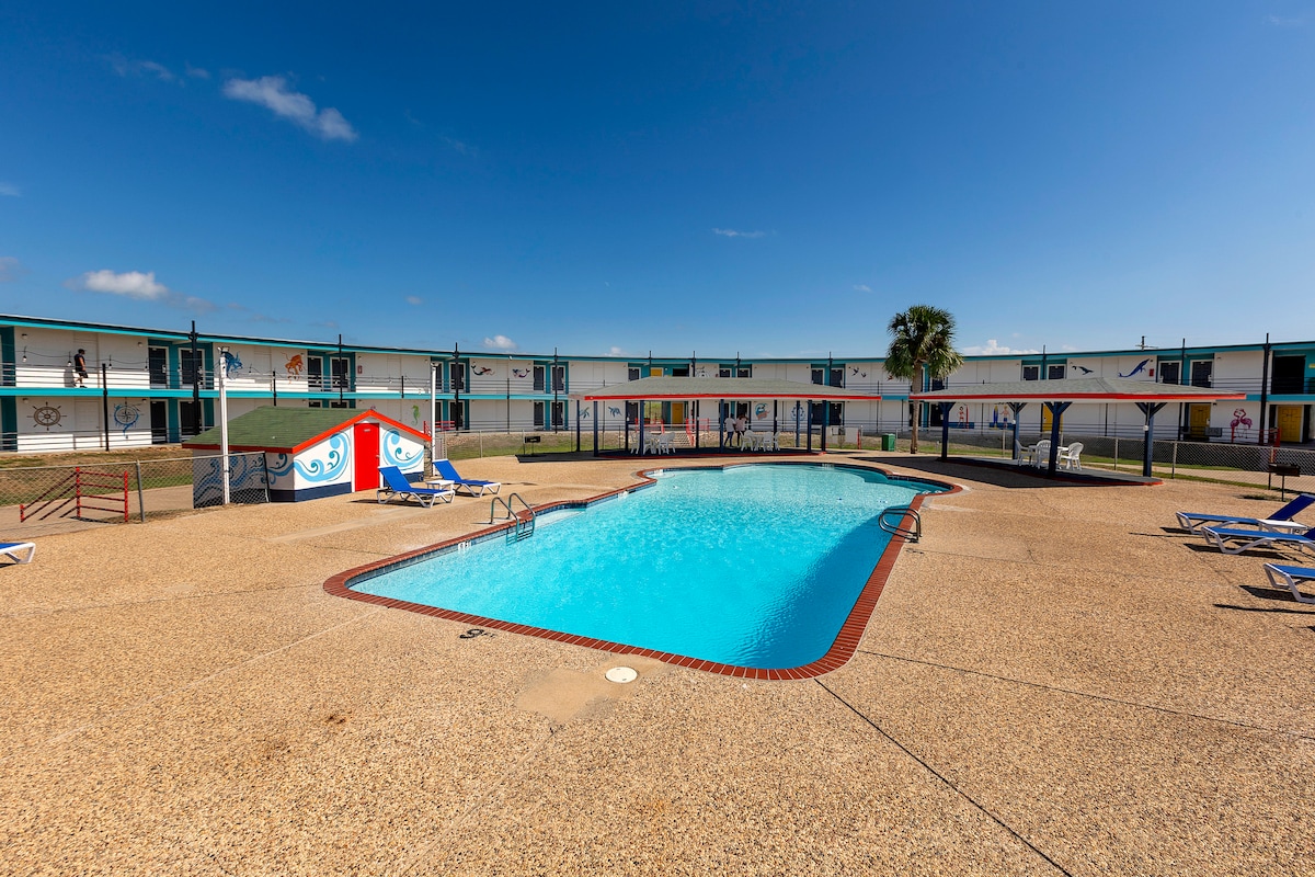 The image features a clear blue swimming pool surrounded by a spacious deck filled with lounge chairs. The pool area is enclosed by colorful murals and two-story guestroom buildings, providing a relaxed outdoor space under a sunny sky.