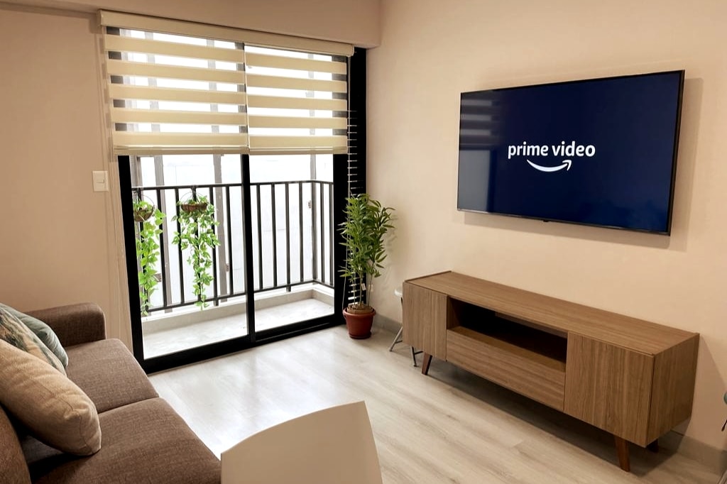 The living area features a modern design, with a comfortable sofa positioned in front of a wooden media console. A 55-inch Smart TV displays the Prime Video logo. Large glass doors provide access to a balcony adorned with potted plants, enhancing the natural light in the room.