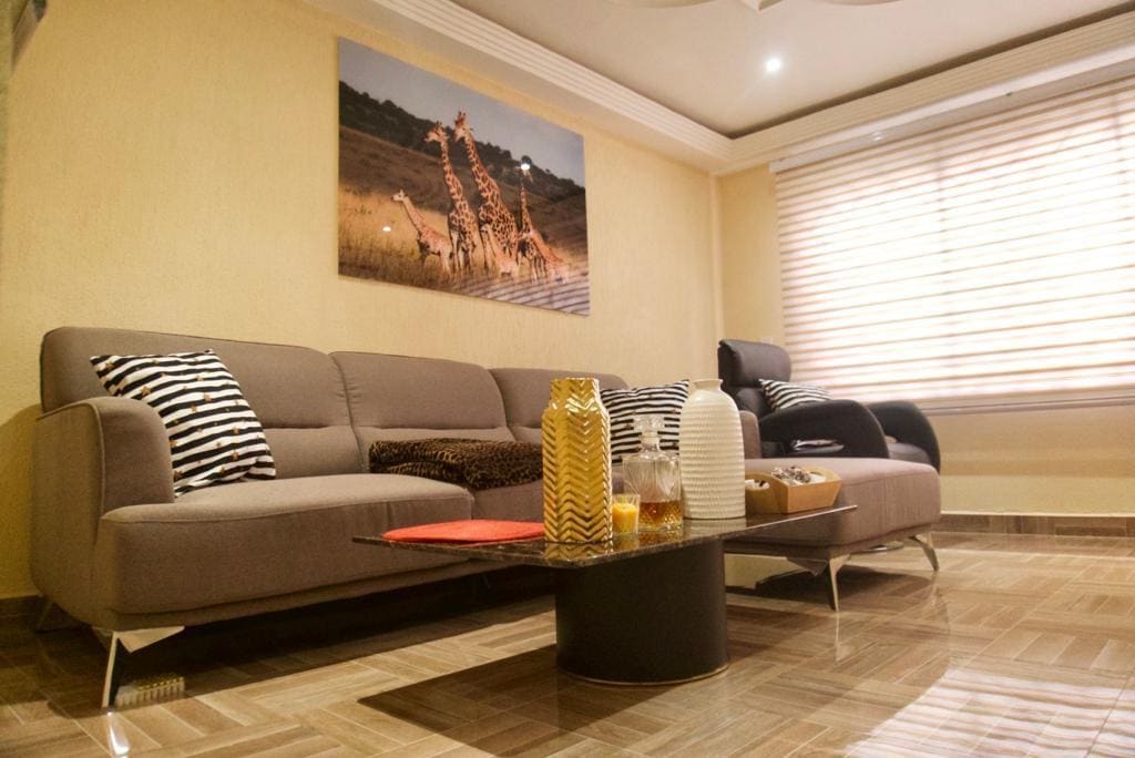 A spacious living area is depicted, featuring modern seating with decorative cushions and a low coffee table. A vibrant artwork of giraffes adorns the wall, and natural light enters through horizontal blinds, illuminating the warm color palette and polished floor.