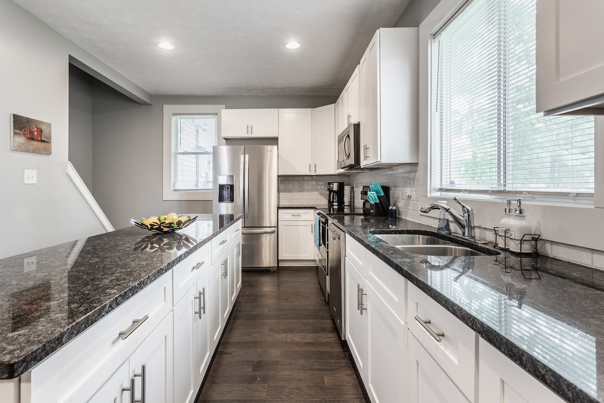 A modern kitchen features sleek white cabinetry and dark granite countertops, complemented by stainless steel appliances. Ample natural light from nearby windows highlights the spacious layout, while a decorative bowl of fruit offers a welcoming touch.