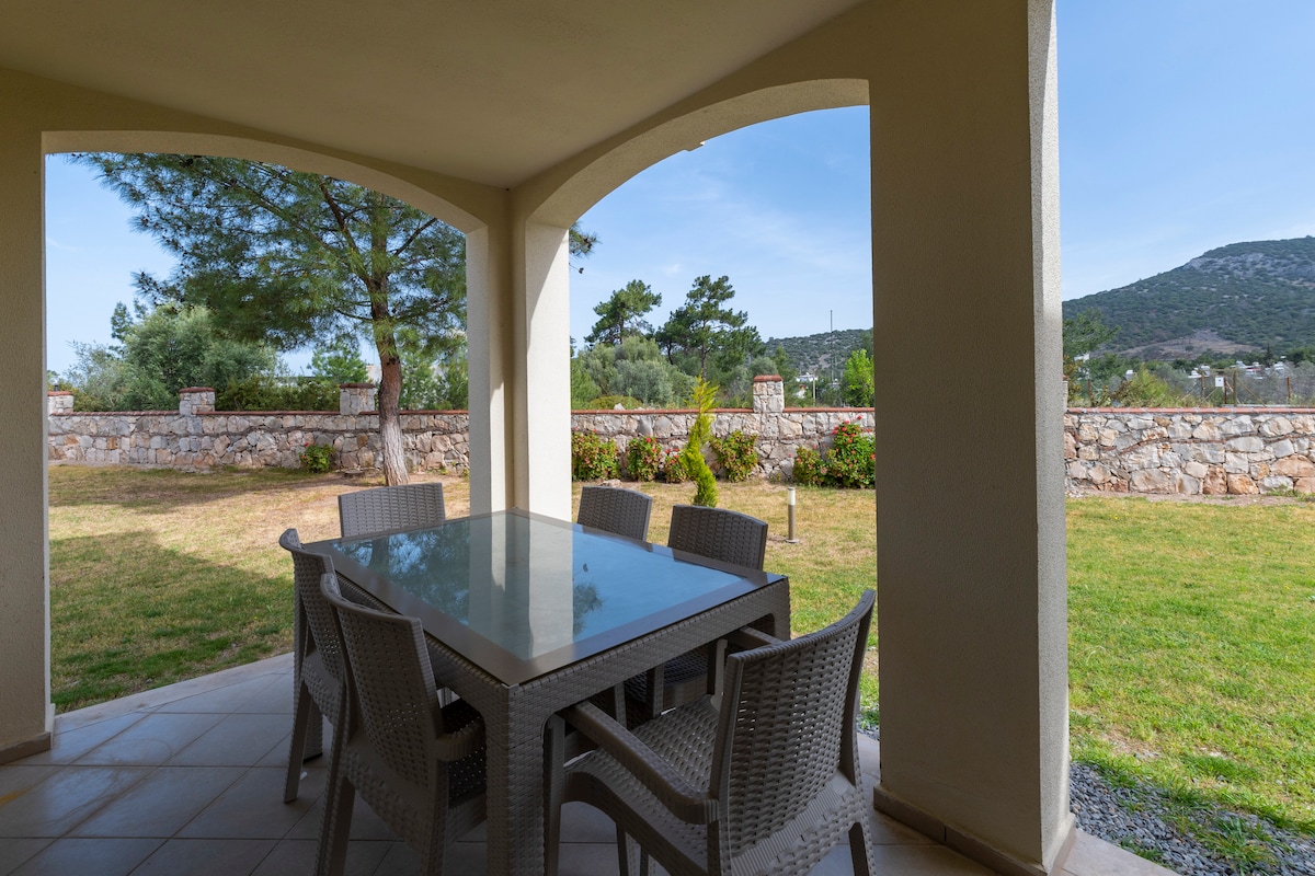 An outdoor patio is visible, featuring a glass-topped table surrounded by six woven chairs. Lush green grass extends around the area, complemented by a stone wall in the background and scattered trees, contributing to a serene outdoor dining experience.