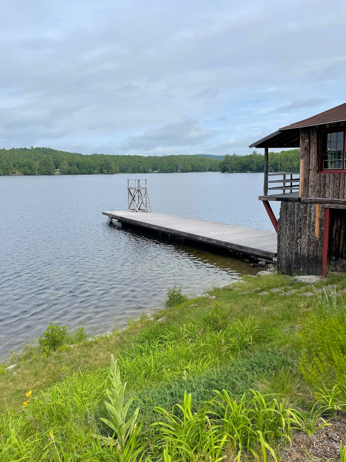 A wooden dock extends into the calm waters of Lovejoy Pond, framed by lush greenery. A rustic cabin is positioned nearby, offering access to the serene waterfront. Soft, cloudy skies reflect over the water, enhancing the tranquil setting.