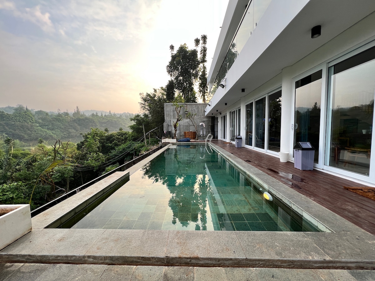 An expansive swimming pool is surrounded by a spacious wooden deck, reflecting the greenery of the landscape. Large glass doors provide a view into the modern living area, and plush outdoor seating is positioned nearby, enhancing the serene outdoor space.