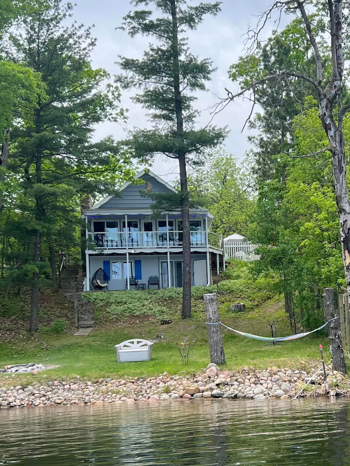 A charming two-story cottage is nestled among trees, featuring a covered porch adorned with seating. The structure overlooks the lake, surrounded by a grassy area with rocky landscaping. A hammock is visible between two trees, creating a relaxing outdoor space.