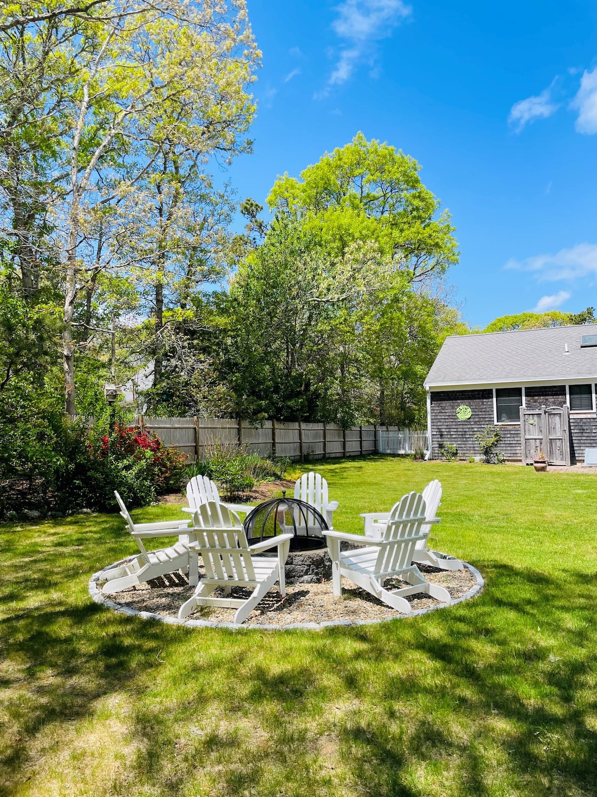 A spacious fenced-in yard is showcased, featuring a circular sitting area with five white Adirondack chairs arranged around a fire pit. Lush greenery and flowering shrubs create a tranquil backdrop, while a home is visible in the distance under a clear blue sky.