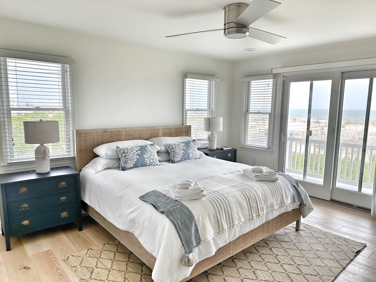 A spacious bedroom features a queen bed with soft linens and decorative pillows. Large sliding glass doors open to a private deck with ocean views. Two nightstands with lamps flank the bed, while natural light fills the room through multiple windows with blinds.