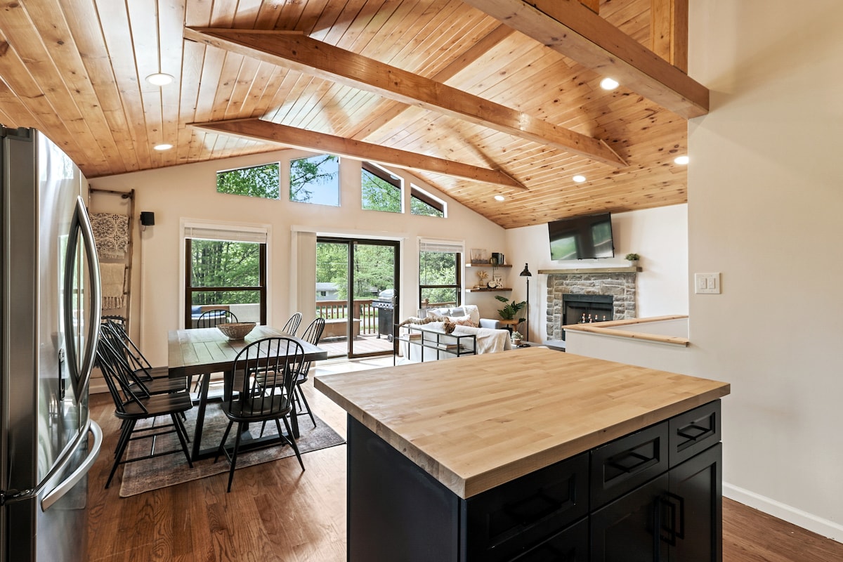 An open-concept living space features a wooden ceiling with exposed beams, large windows, and sliding glass doors that provide natural light and views of the outdoors. A dining table with black chairs is positioned next to a kitchen island with a light wood surface.