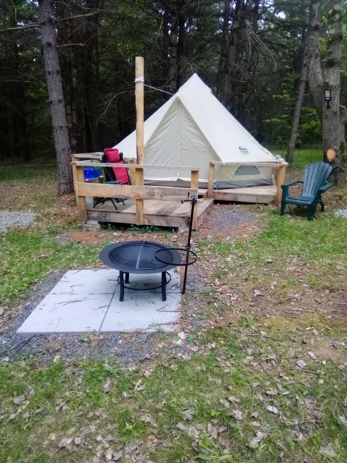 A camping area is depicted with a conical tent situated on a wooden platform. Surrounding the tent, a fire pit is placed on a stone surface, and a green Adirondack chair is positioned nearby, complemented by scattered leaves and natural vegetation.
