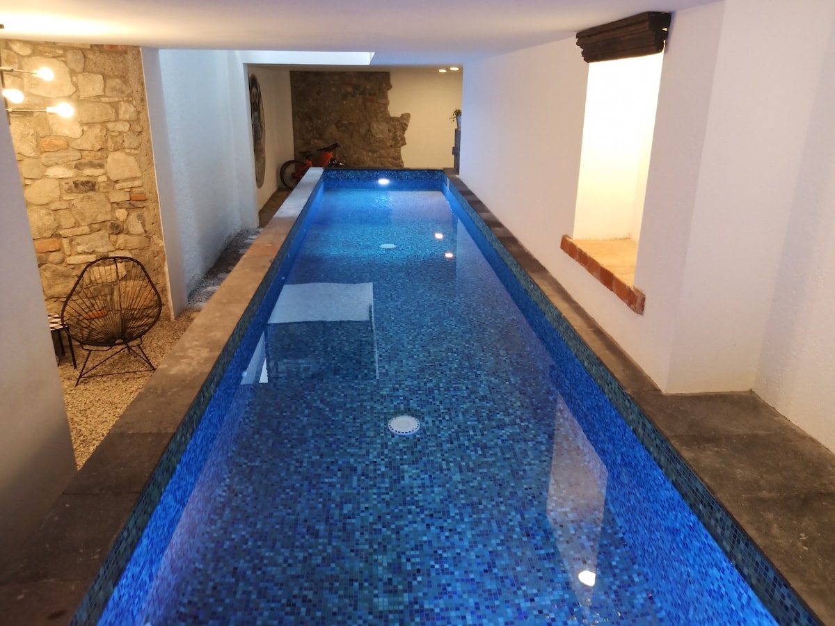 A narrow, indoor swimming lane is depicted, featuring clear blue waters and a mosaic tile finish. Soft, ambient lighting highlights the space. A black, woven chair is positioned nearby, adding a touch of modern comfort to the serene environment.