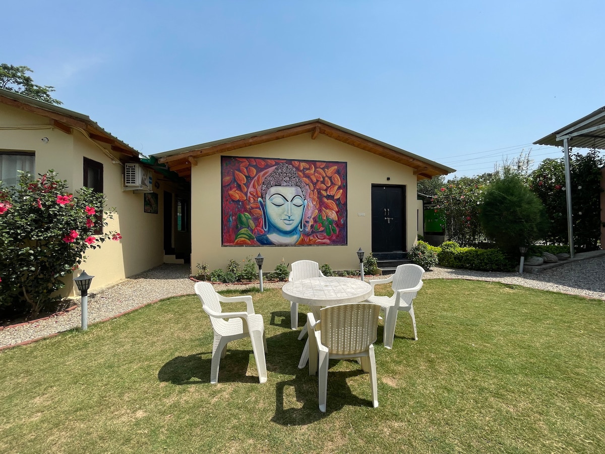 An outdoor area features a circular table surrounded by four white chairs, situated on a grassy lawn. A colorful mural decorates the wall of a nearby building, framed by manicured hedges and blossoming flowers. Clear blue skies provide a bright backdrop.