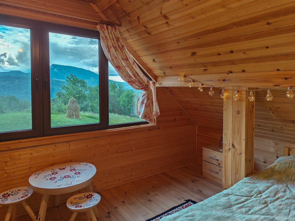 A cozy bedroom features wooden walls and a sloped ceiling, enhanced by a large window offering views of the surrounding mountains and fields. A small table with patterned design sits beside the bed, illuminated by soft lighting from decorative bulbs.