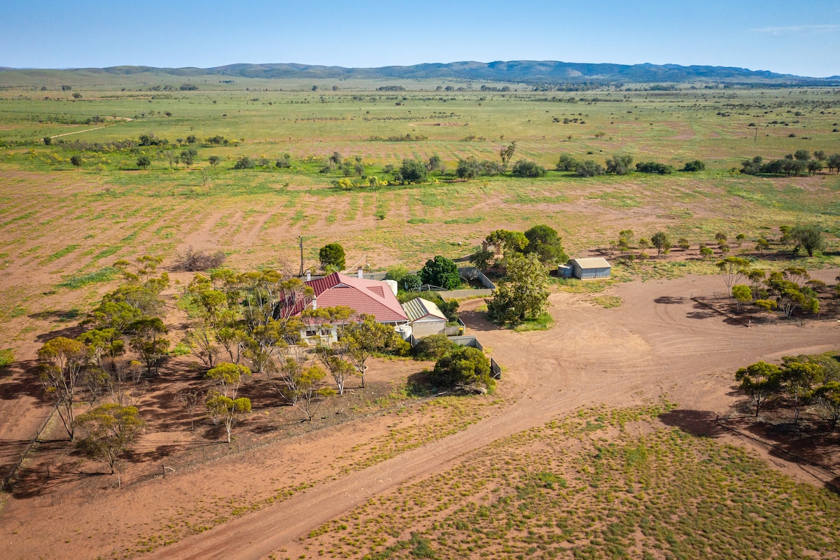 An aerial view captures Crotta Homestead amid expansive, undulating landscapes. The homestead features a red roof and is surrounded by sparse trees, with dirt paths leading to nearby structures. The open surroundings highlight the vastness of the property, showcasing its rural setting.