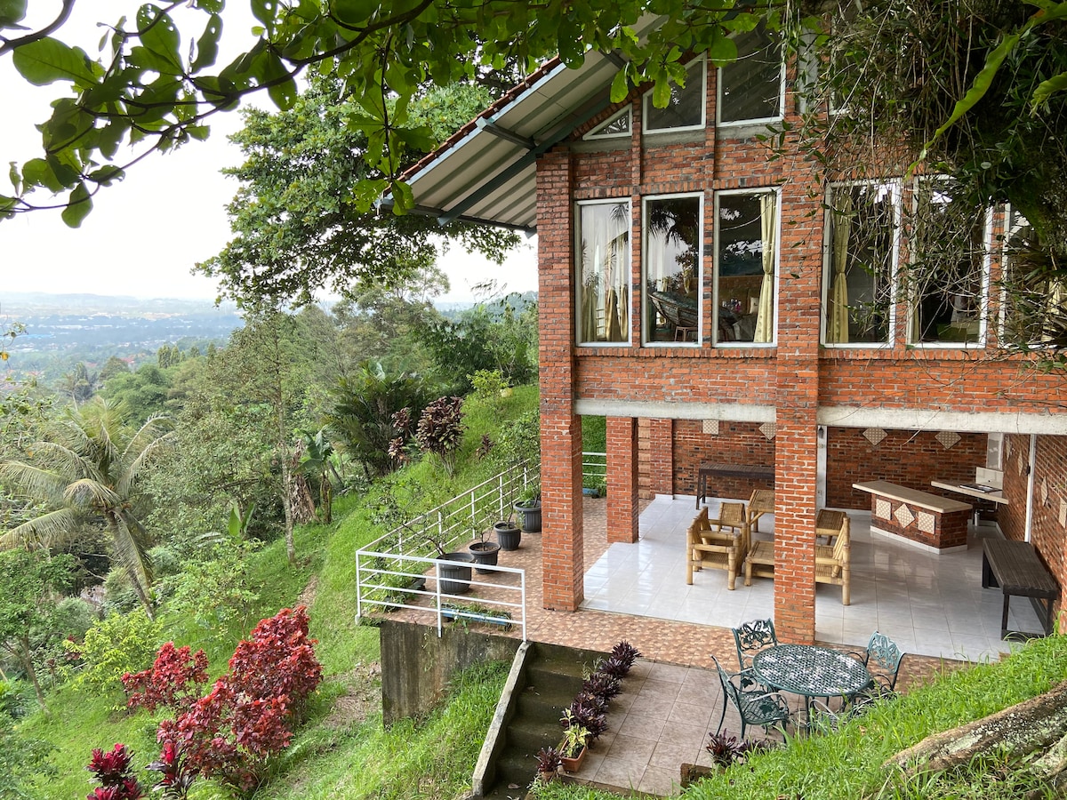 A modern villa is situated on a hillside, surrounded by lush greenery. Large glass windows allow natural light to fill the interior. The outdoor area features a dining table and chairs on a tiled patio, overlooking a vibrant landscape.