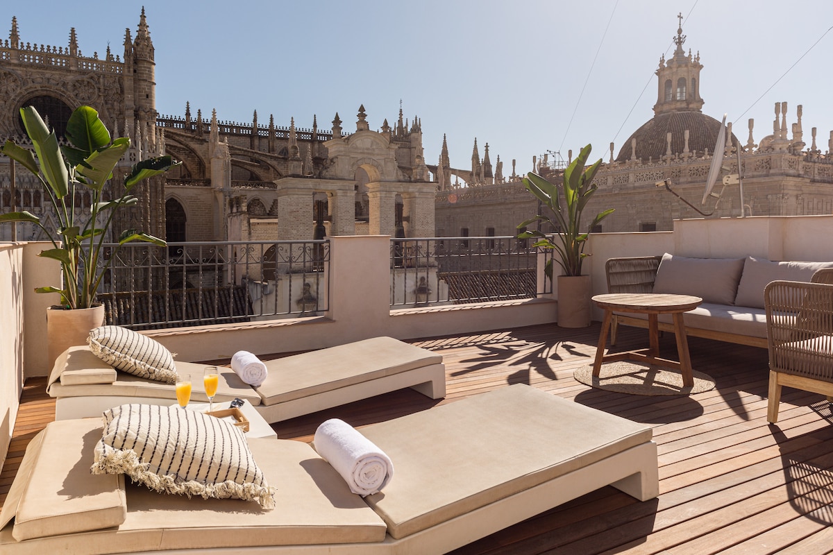 A private rooftop terrace features two sun loungers with plush cushions, accompanied by a small round table set with two glasses of juice. Lush green plants are placed around the area, providing a serene atmosphere. The stunning view showcases the Cathedral and its intricate architecture.