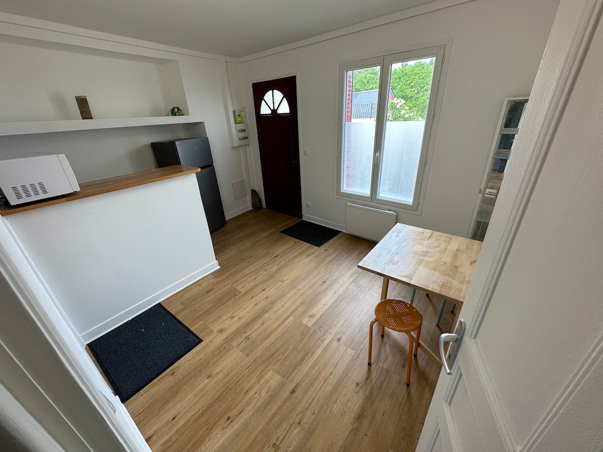 An inviting entryway features warm wooden flooring and a simple wooden table with a chair. A small kitchenette area is visible with a countertop and refrigerator. Natural light floods in through a large window, enhancing the bright and open atmosphere.