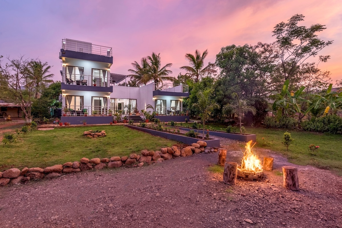 A modern villa is showcased against a twilight sky, framed by lush greenery and palm trees. A bonfire burns in a circular stone pit surrounded by wooden logs, adding warmth to the serene outdoor setting. The expansive lawn and well-maintained garden complement the structure.