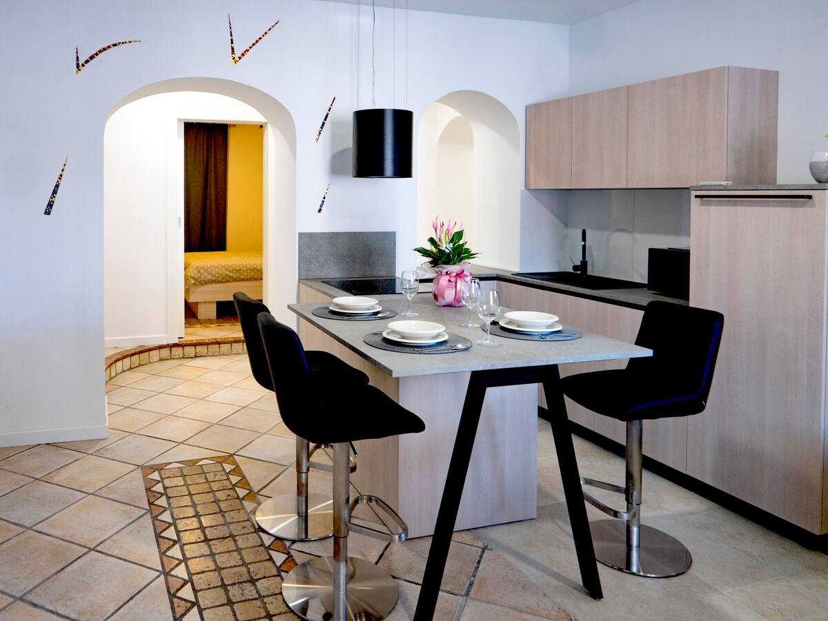 A modern kitchen area features a light-colored table with four high-backed stools, accompanied by a neutral-toned countertop. Arches lead to a separate room in the background, while decorative elements adorn the walls, adding character to the open space.