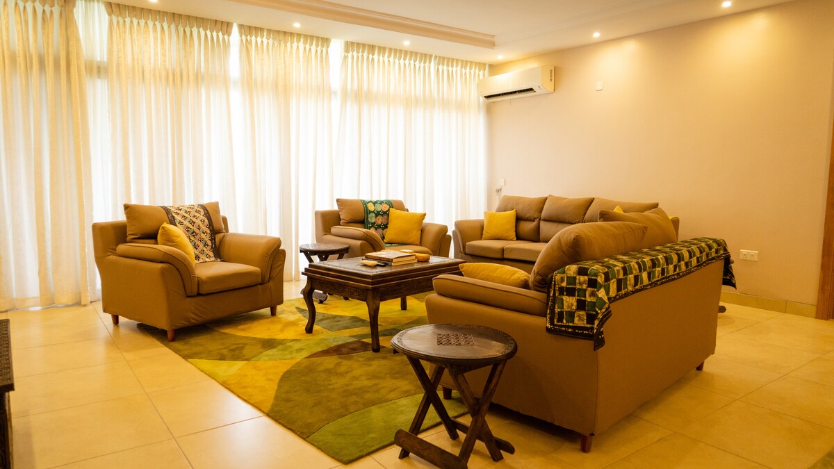 A bright living area features comfortable seating arranged around a wooden coffee table. Light-colored curtains drape over large windows, allowing natural light to fill the space. A patterned rug adds a touch of color to the tiled floor, complementing the neutral tones of the furniture.