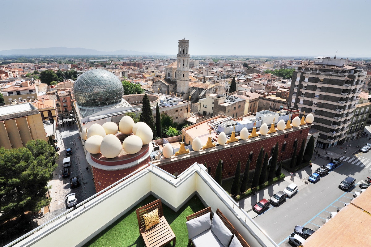 A panoramic view from a private terrace reveals the urban landscape, including notable landmarks such as the Torre Galatea and the church of Sant Pere. The scene encompasses greenery on the terrace, accentuated by lounge furniture, with a clear sky above and distant mountains on the horizon.