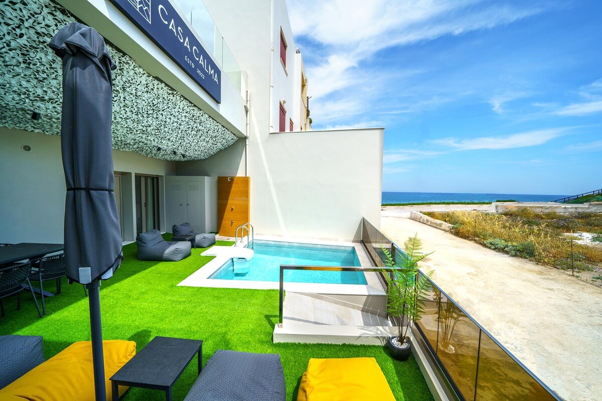 An outdoor area features a private pool with steps leading into the water. Six comfortable bean bags surround the pool, while a large table with chairs provides a dining option. A sun umbrella stands nearby for shade, with views of the sea beyond the property.