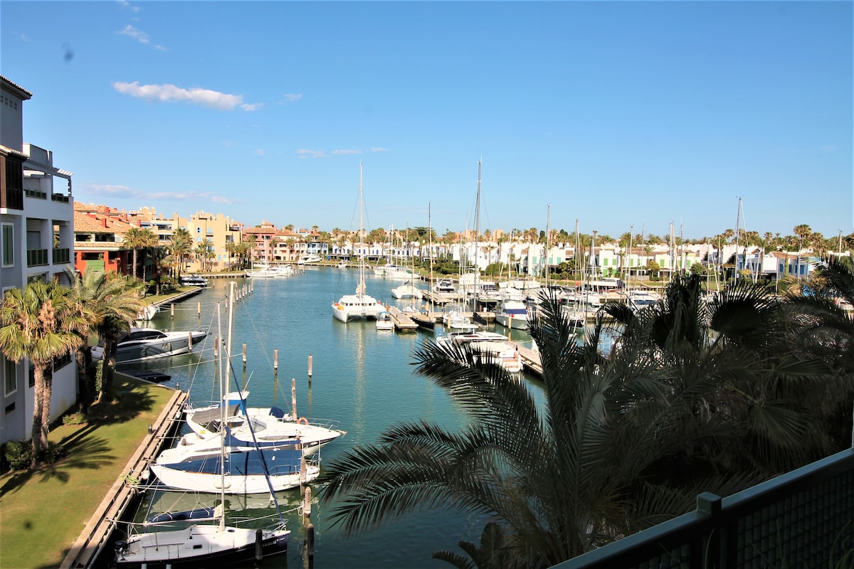A scenic view of a peaceful marina is captured, featuring various boats moored along the calm water. Lush palm trees line the waterfront, with colorful buildings visible in the background, blending harmoniously with the vibrant landscape.