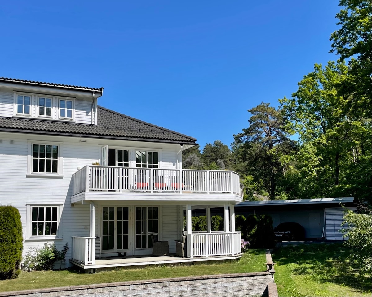 The exterior of a charming white two-story building is displayed, featuring a spacious balcony with seating. A carport is visible beside the structure, surrounded by greenery and trees, against a clear blue sky.