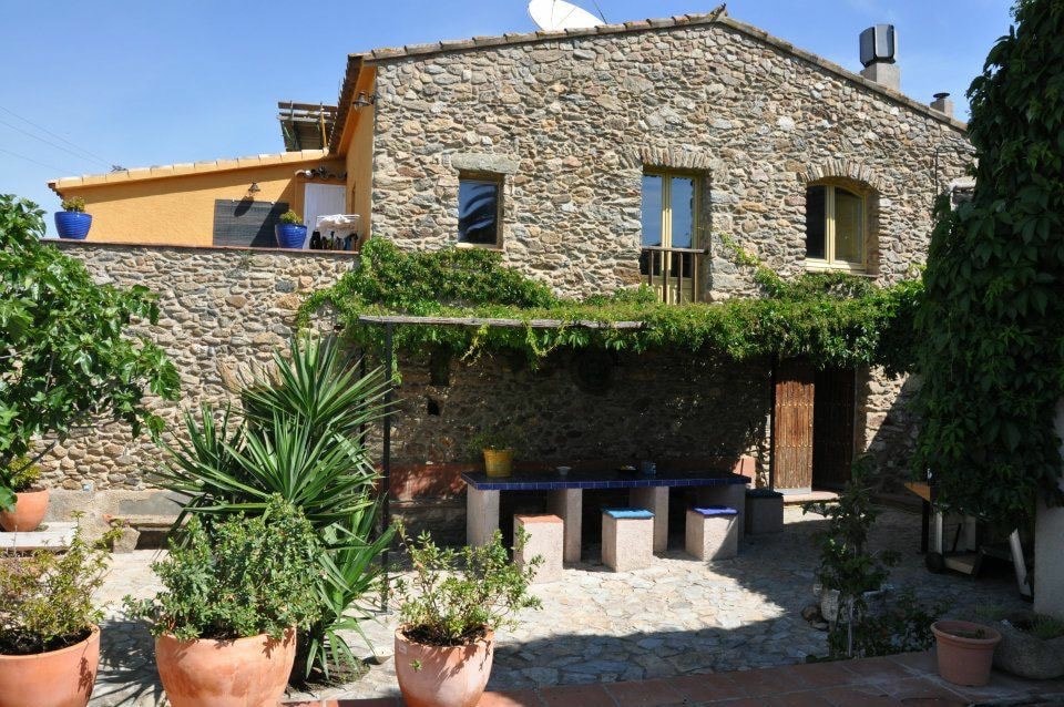 The exterior of the stone masía is framed by lush greenery and potted plants. A quaint courtyard with stone flooring is present, featuring several seating areas against the building. Windows are adorned with flower boxes, creating a welcoming outdoor space.