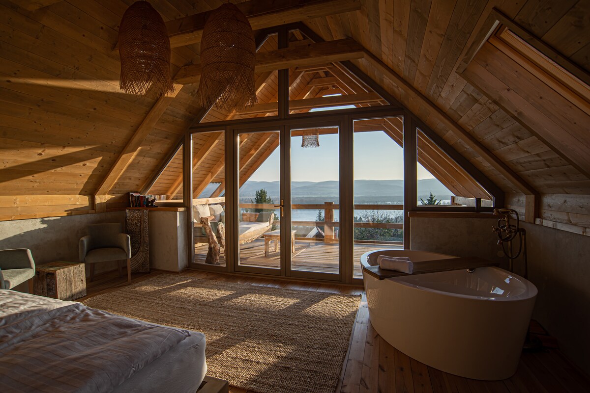 The spacious attic room features a modern freestanding bathtub and a large window with panoramic views of the landscape. Natural materials are used throughout, with warm wooden beams and a soft area rug complementing the décor. A comfortable seating area is visible near the window.
