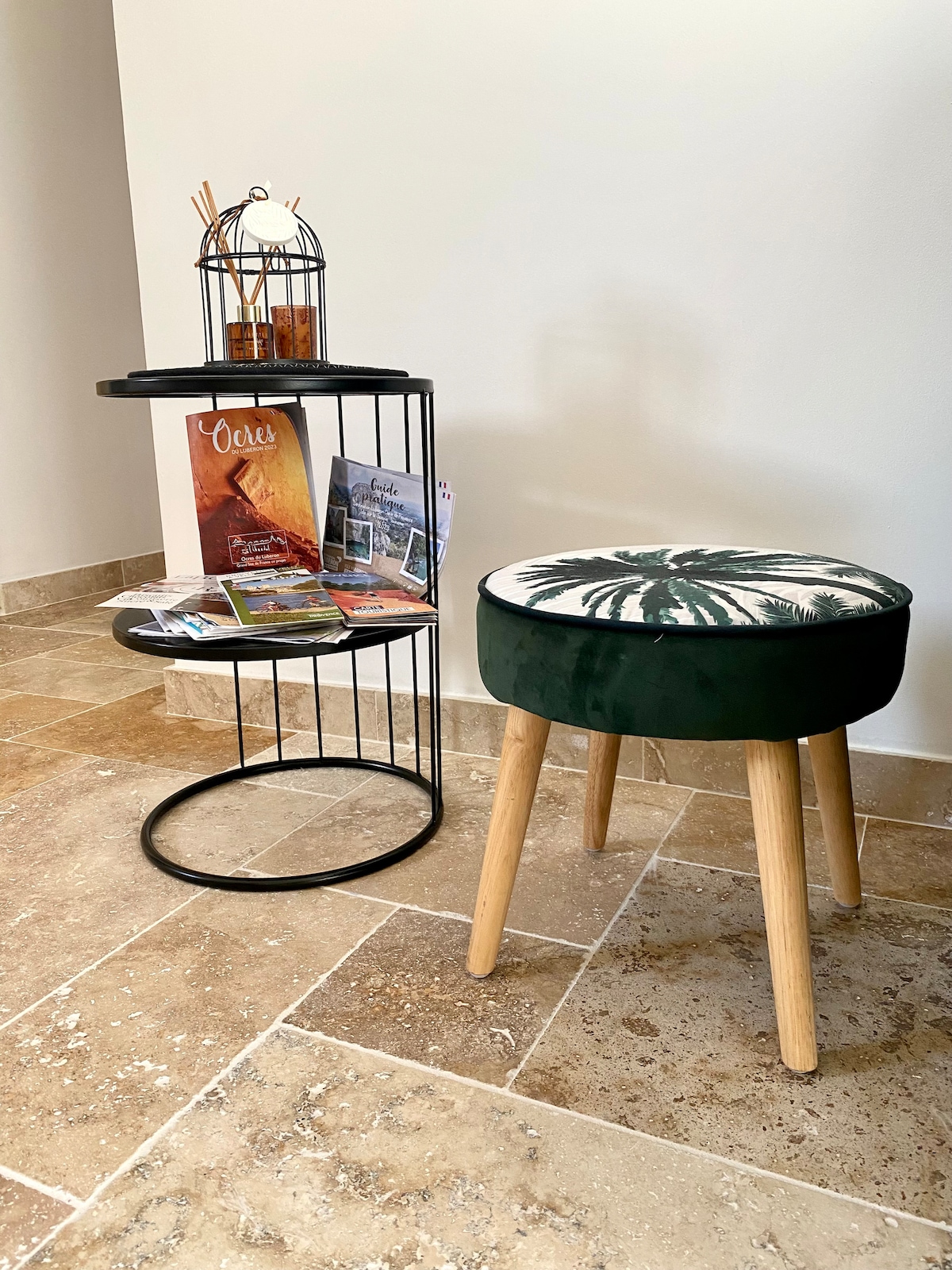A modern side table with two round shelves is positioned beside a green upholstered stool. The lower shelf displays brochures and travel materials. The flooring features natural stone tiles, adding texture to the neutral-toned space.
