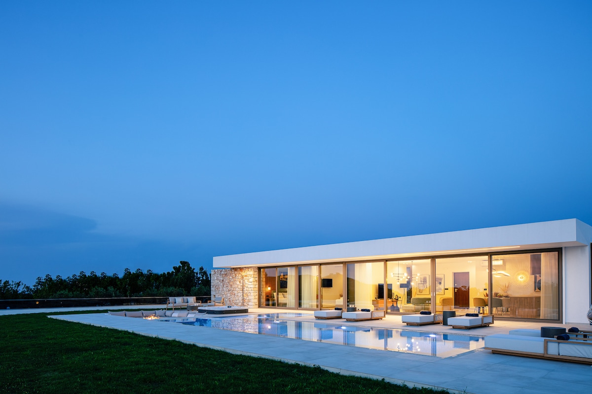 A modern villa is illustrated under a twilight sky, featuring large glass doors that provide a seamless connection to the outdoor space. The private swimming pool reflects soft light, while lounge areas are visible around the pool, set on a lush green lawn.