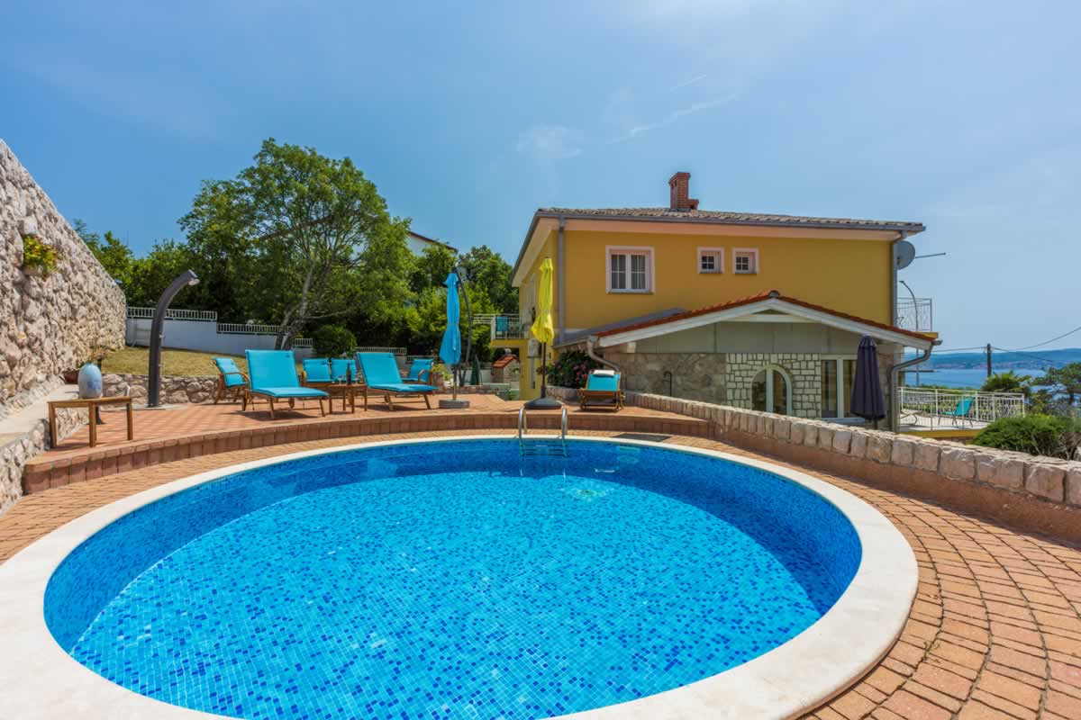 A circular swimming pool is surrounded by a brick patio and several sun loungers, offering a relaxing spot for guests. The property features a two-story building in the background, framed by greenery and a clear sky.