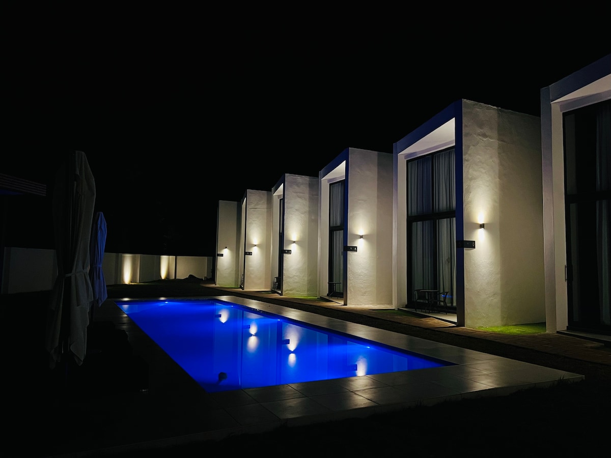 An exterior view of modern accommodations is presented at night, featuring illuminated walls and a tranquil swimming pool. Soft lighting reflects off the water, surrounded by clean lines of the building and a peaceful garden area.