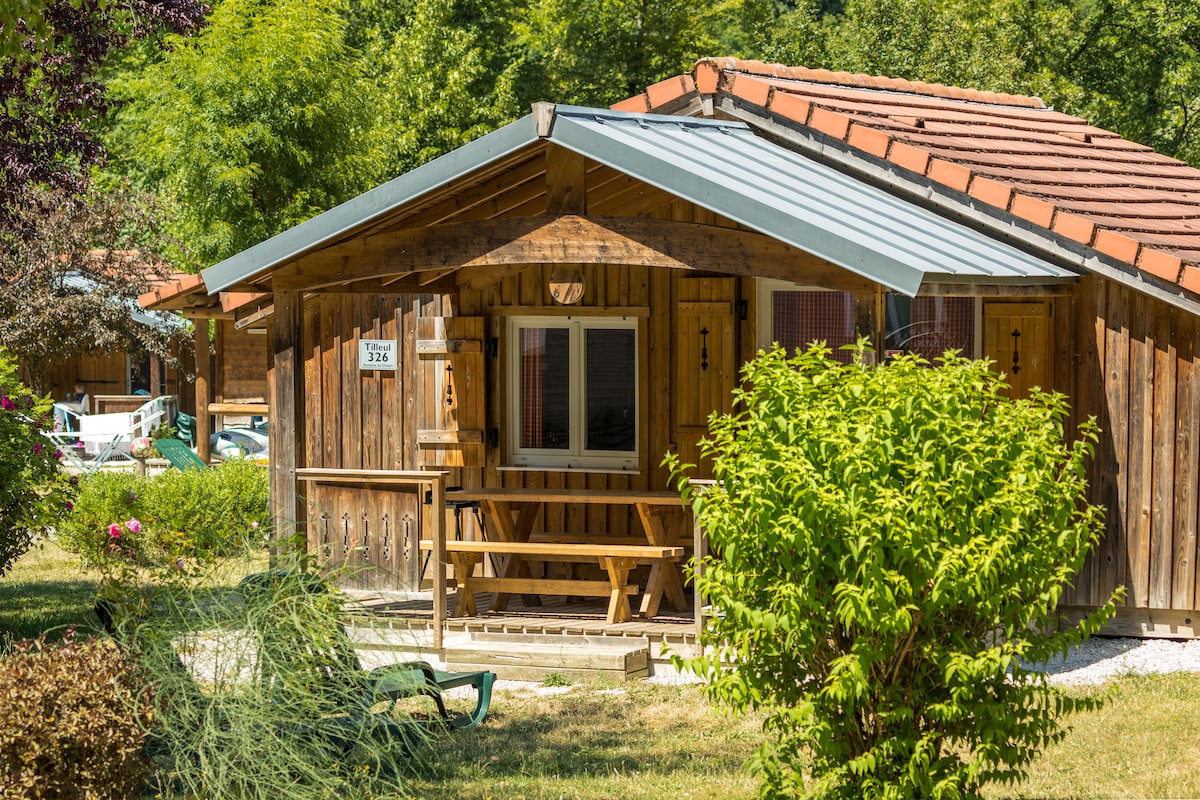 A wooden chalet features a sloped roof with metal accents, blending into the natural surroundings. A small covered porch is furnished with a bench, offering a welcoming entrance. Lush greenery and flowering bushes frame the chalet, enhancing the outdoor ambiance.