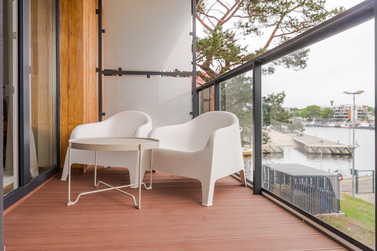 A furnished balcony features two white chairs and a circular table, providing a relaxing space. Large glass doors lead to open views of the marina and surrounding landscape, allowing natural light to enhance the ambiance.