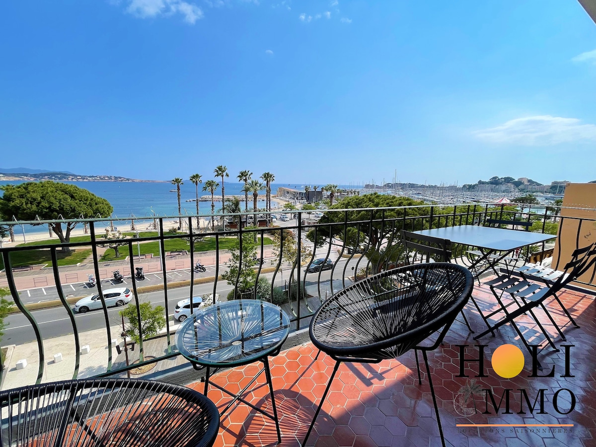 A balcony view overlooking the coastline, showcasing a wide expanse of the sea and beach. Two black outdoor chairs are positioned around a small table, framed by palm trees and the harbor in the distance. Clear blue skies complement the vibrant scenery.