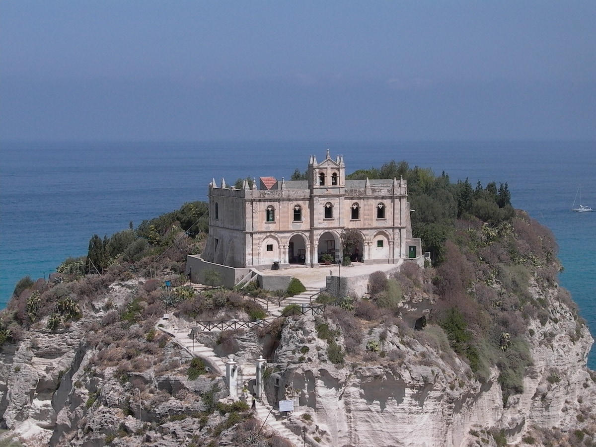 A historic building is positioned on a rocky cliff, overlooking a calm sea. Shaded by surrounding greenery, the structure features multiple arches and a distinctive rooftop. Clear skies and gentle waves contribute to a serene coastal setting.