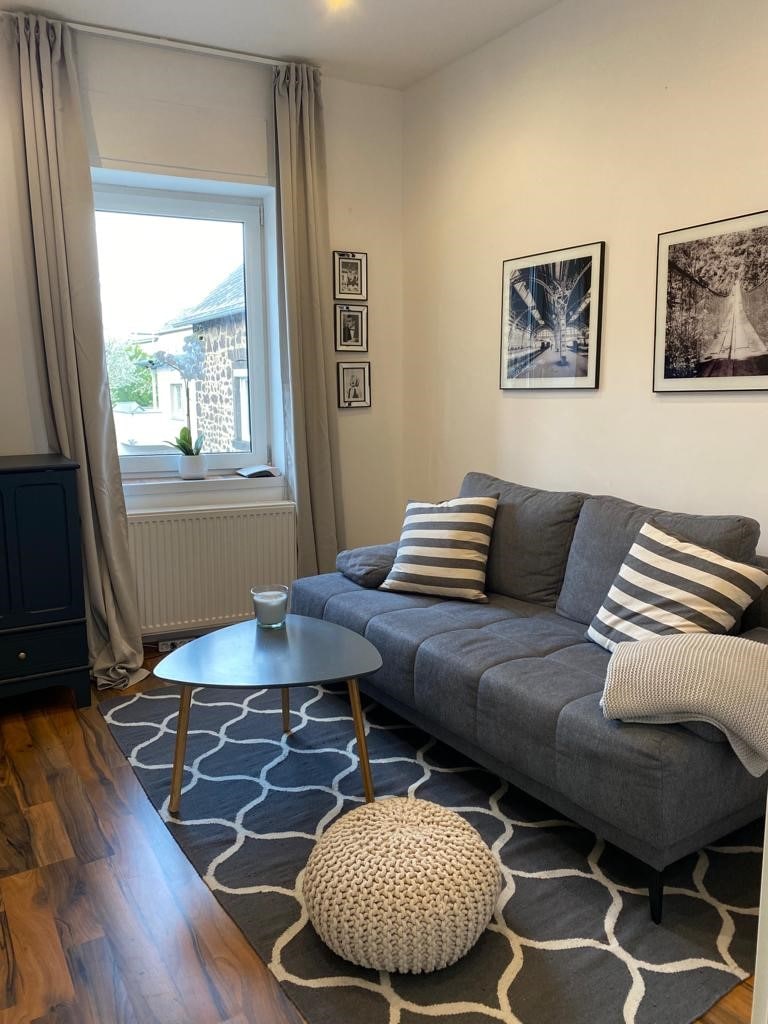 A cozy living room is showcased, featuring a grey sofa adorned with striped cushions. A round coffee table is positioned in front, accompanied by a textured pouf. Two framed photographs are hung on the wall near a window, which is draped with soft curtains.