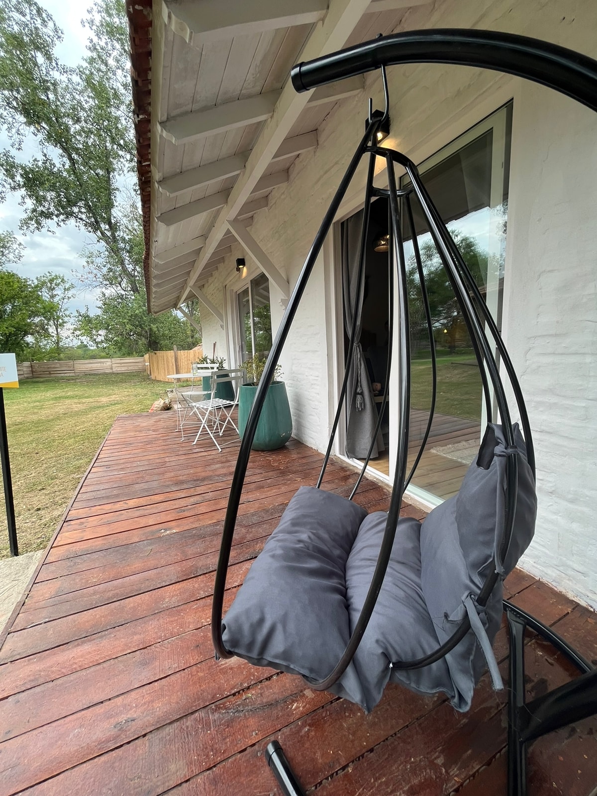 A suspended swing chair with soft gray cushions is positioned on a wooden deck. The backdrop features a well-kept lawn and a quaint outdoor seating arrangement, creating a relaxing space to enjoy nature. Large windows provide a view of the surrounding area.