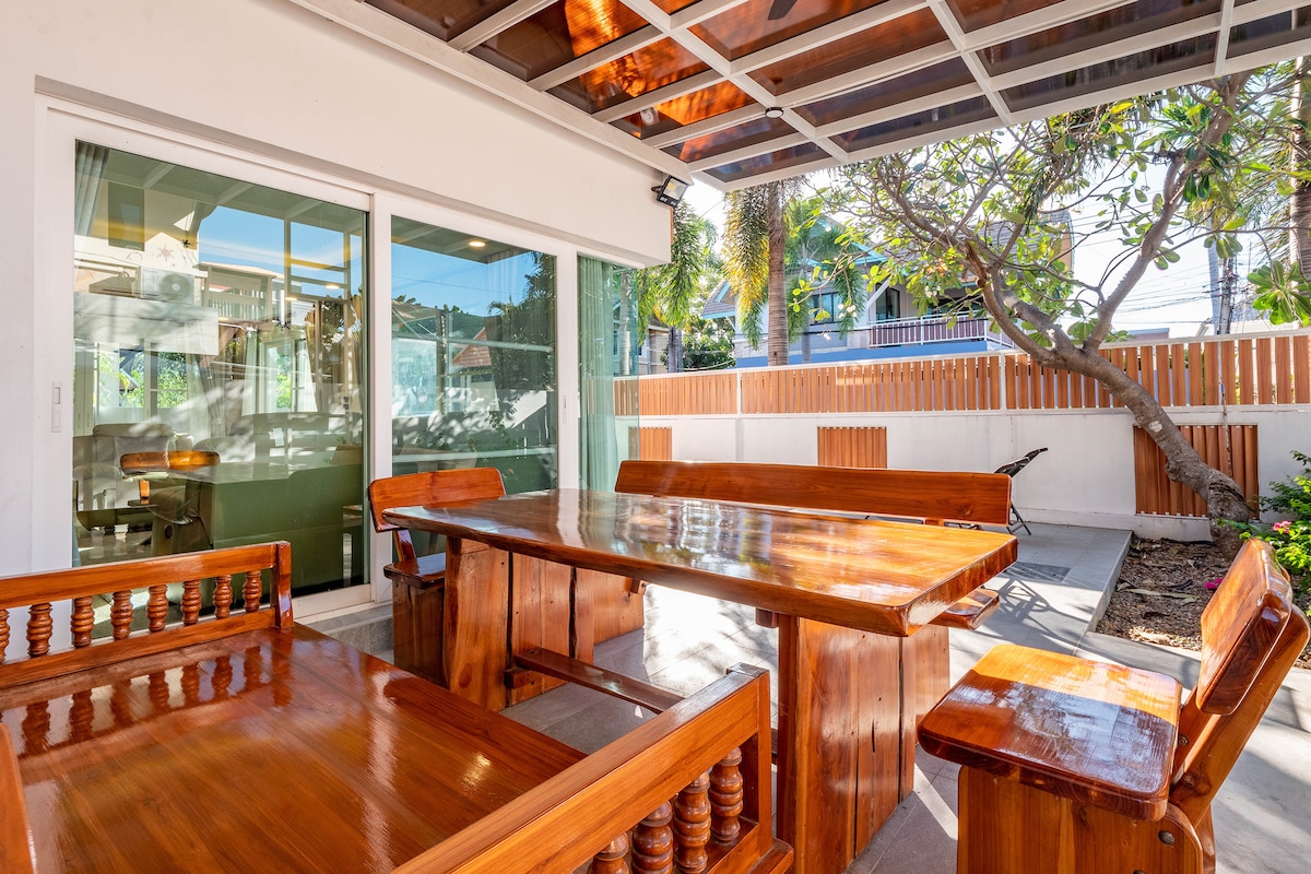 A spacious outdoor dining area features a large wooden table surrounded by sturdy wooden benches. Sunlight filters through a glass canopy, illuminating the setting. Lush greenery is visible in the background, enhancing the inviting outdoor experience.