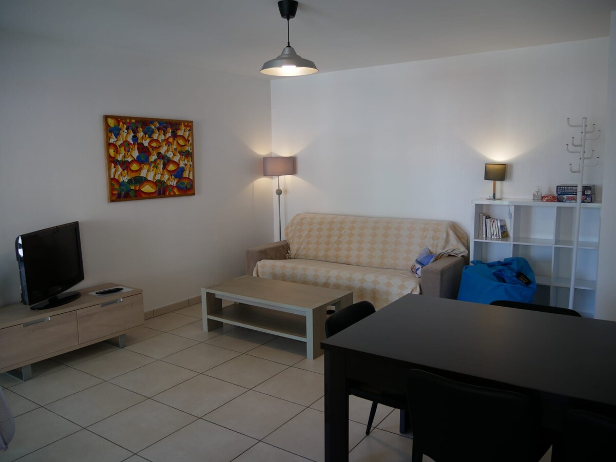 A living area is visible, featuring a light-colored sofa covered with a patterned throw. A coffee table sits in front, and a TV is positioned on a low cabinet. Additional lamps provide soft lighting, while a colorful wall art piece adds visual interest. A dining table is seen in the background.