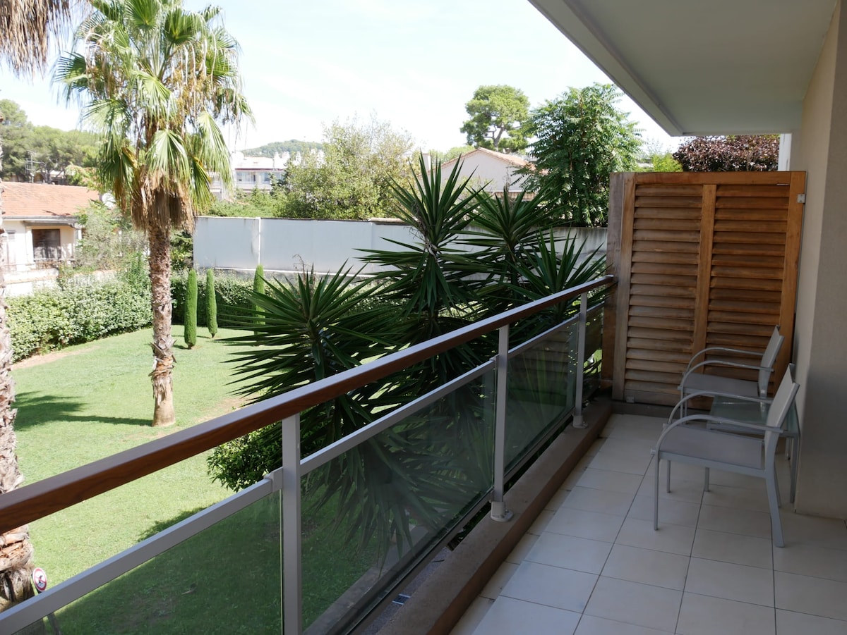A spacious balcony is presented, featuring two chairs positioned against a railing. Lush greenery is visible in the surrounding landscape, including palm trees and shrubs. The area appears tranquil, offering a view of the well-maintained garden below.