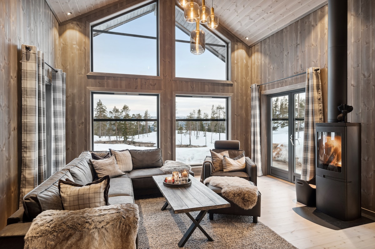 The living area features large windows allowing natural light to fill the space, offering views of the snowy landscape. A cozy sectional sofa is arranged around a coffee table, with warm textiles enhancing the ambiance. A modern fireplace adds a focal point to the room.