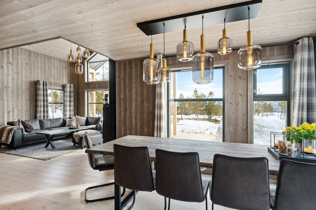 A modern dining area features a sleek table with five dark chairs and a pendant light above. Large windows provide views of snow-covered trees, with natural light illuminating the wooden walls. A comfortable seating area is visible in the background.