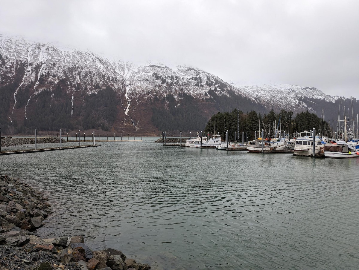 A calm harbor is depicted with several boats docked along wooden piers. Snow-capped mountains rise in the background, partially shrouded by a gray sky. The water reflects the surroundings, creating a serene atmosphere.