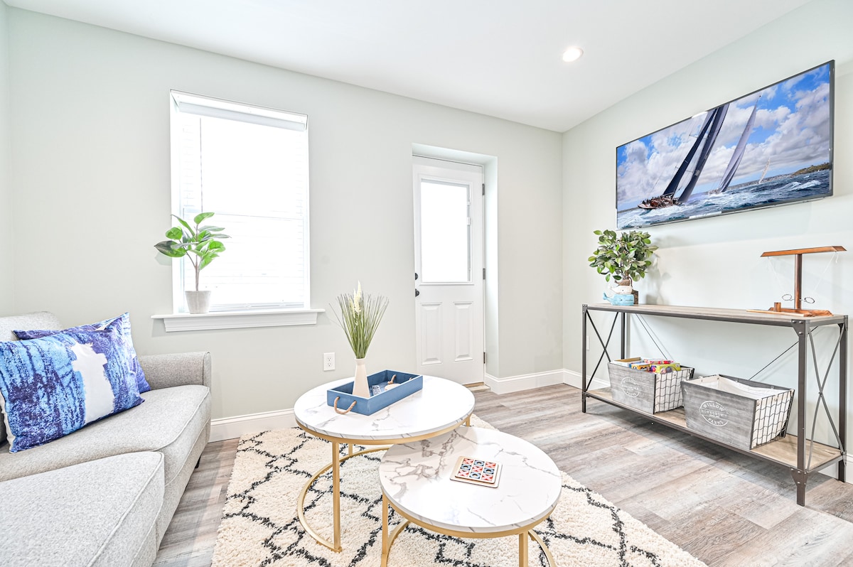 A light-filled living area is presented with a comfortable gray sofa and two round coffee tables. A lush indoor plant adds a touch of greenery, while a large television is mounted on the wall, displaying a nautical scene. A decorative shelving unit holds small baskets.
